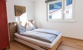 A cozy bedroom with a double bed and checkered pillows. The room has light walls, a large window, and a red cabinet.