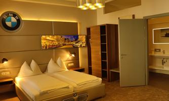 A modern bedroom with two single beds and a BMW logo on the wall. The room is bright and stylishly furnished, with a small desk and an open wardrobe.