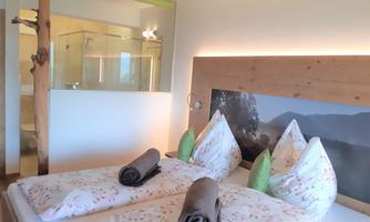 A cozy bedroom with two beds and fresh flower bedding. In the background, a modern bathroom with a glass shower cabin can be seen.