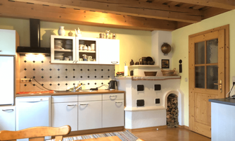 A cozy kitchen with white furniture and wooden ceilings. Next to the stove is a fireplace with a wood storage.