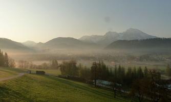 A picturesque landscape with gentle hills and mountains in the background. The sky is clear and the morning mist gives the scene a tranquil atmosphere.
