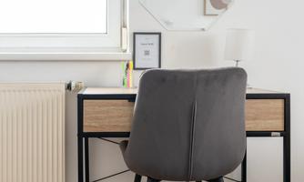 A modern desk with a gray chair is located in a bright room. Next to the table is a window and a decorative wall mirror.