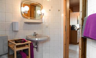 A modern bathroom with white tiles and a small sink. In the foreground is a purple bath mat, and there is a wooden shelf with towels.