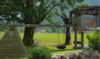 A beautiful garden with a swing and a wooden playground under a large tree. In the background, fields and mountains are visible.