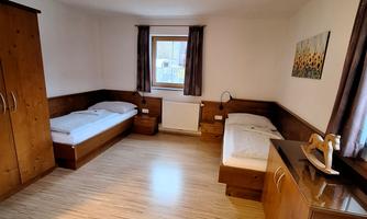 A bright room with two single beds and a large window. The walls are painted in neutral colors, and there is a comfortable wooden wardrobe.