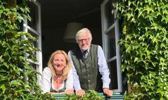A couple is standing smiling at the window, surrounded by green ivy. The atmosphere is friendly and inviting.