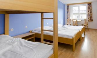 A simple room with two beds and a bunk bed. There is a table with chairs and large windows that let in natural light.
