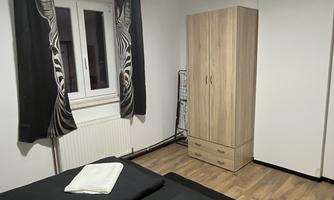A simple bedroom with two beds and a wardrobe. The walls are light, and there are large windows with black curtains.