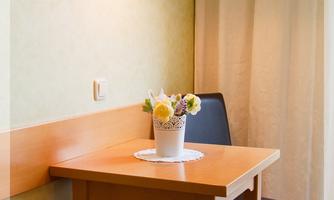 A cozy table with a small flower vase is in a bright room. Next to the table is a black chair and a warm white lamp on the wall.