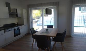 A modern kitchen with a dining area. The table is equipped with chairs and there are large windows that let in light.