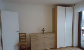 A bright room with wooden flooring, a wardrobe, and a chest of drawers. A lamp and a chair are also available.