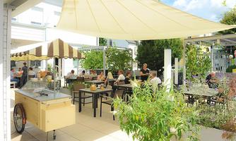 An inviting café with outdoor tables and sun umbrellas. Many guests are enjoying their time in a green environment.