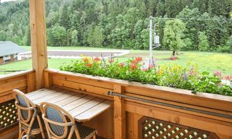 A balcony with a wooden table and chairs, surrounded by blooming plants. In the background, green forests and a meadow can be seen.