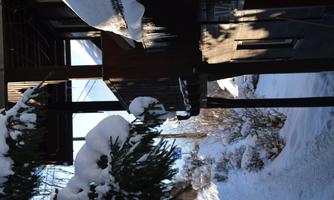 A snow-covered village with a wooden house and a balcony. The surroundings are wintry with icy branches and a narrow walkway.