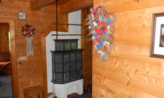 A cozy hallway with wooden walls and a traditional tiled stove. On the wall, a floral wreath and a picture frame can be seen.