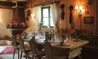 A cozy dining area with a large table and wooden chairs. In the background, a fireplace flickers, and the walls are decorated in a rustic style.