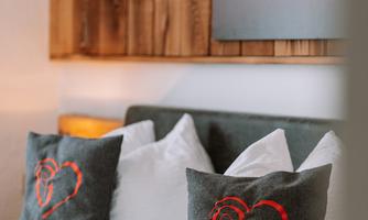 A modern bedroom with cozy bedding and heart-patterned pillows. A wooden panel in the background gives the room a warm atmosphere.