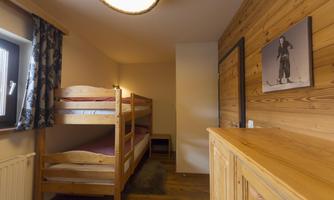 A simple bedroom with a loft bed and a wooden wardrobe. The walls are made of wood and there is a window with curtains.
