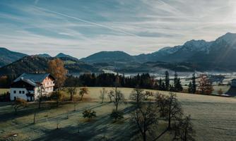 A picturesque landscape with a farmhouse, surrounded by green meadows and trees. In the background, majestic mountains and a clear sky are visible.