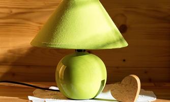 A green table lamp with a round base and a slanted shade is standing on a table. Next to it lies a heart-shaped wooden object.