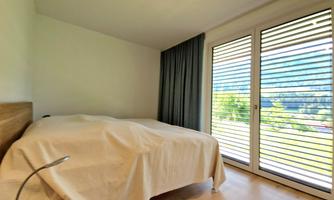 A bright bedroom with a bed and large windows. Through the windows, you can see a green landscape and a clear sky.