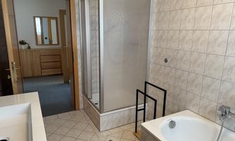 A modern bathroom with a shower and a bathtub. The walls are tiled in light colors, and in the background, you can see a wooden cabinet.