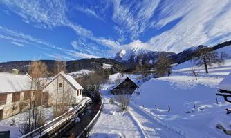 A snowy landscape with houses and gentle hills. The sky is clear and blue with impressive cloud formations.