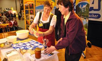 Two women are standing at a table, preparing a dish together. In the background, colorful posters and a crowd of people can be seen.