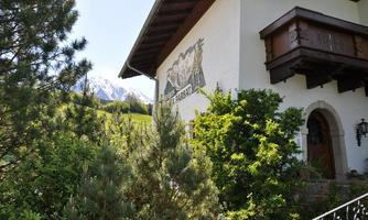 A charming house with a balcony and a picturesque facade. In the background, snow-capped mountains and lush greenery can be seen.