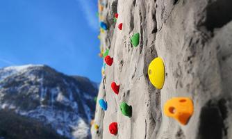 A climbing wall with colorful grips in a wintry landscape background. In the background, snow-covered mountains and a blue sky are visible.