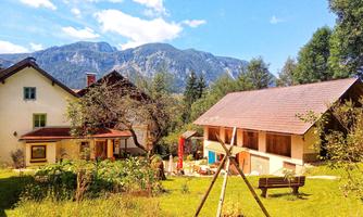 An idyllic landscape with two houses and a green meadow. In the background, mountains and a blue sky can be seen.