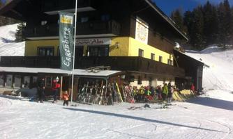 A ski lodge with a yellow facade is situated in a snow-covered landscape. Various skis are leaning against the wall, and guests are enjoying the sun.