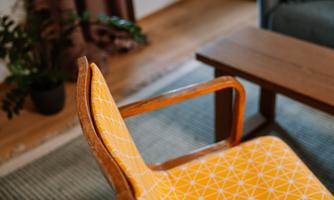 An elegant chair with orange, geometric upholstery stands in a cozy room. The floor is made of wood, and there is a table as well as plants in the background.