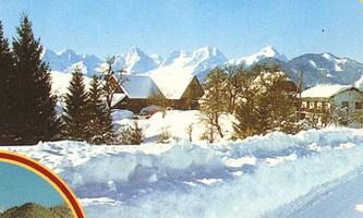 A snowy landscape with huts and high mountains in the background. The trees are covered in snow, and the sky is clear.