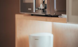 A modern microwave is placed on a shelf above a coffee maker and a kettle. The appliances are situated in a bright kitchen environment.