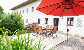 An inviting outdoor area with a table and chairs under a red sunshade. Surrounded by greenery and gravel, this spot offers a pleasant atmosphere for relaxation.