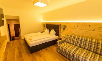 A modern hotel room with a double bed and a cozy couch. The room is designed in warm colors and features wooden paneling.
