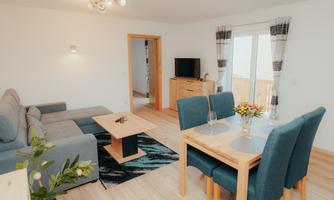 A modern living and dining room with a grey sofa and a wooden table. Large windows let in plenty of light and provide a cozy setting for relaxation.