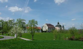 A picturesque landscape with gentle hills, green meadows, and blooming trees. In the background, a historic building with domes can be seen under a clear blue sky.
