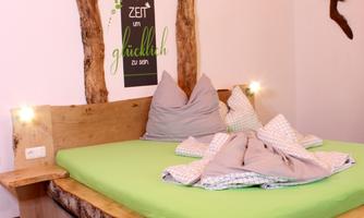 A cozy bedroom with a large wooden bed and a green duvet. The walls are light and decorated with decorative details.