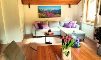 A cozy living room with a bright sofa, colorful cushions, and a large window. Fresh tulips are in a vase on the table.