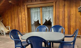 A table with six blue chairs stands on a wooden terrace. In the background, there is a window with white curtains and a wooden facade.