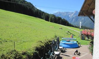 A green meadow with mountains in the background and a clear blue sky. In the foreground, a swimming pool and seating can be seen.