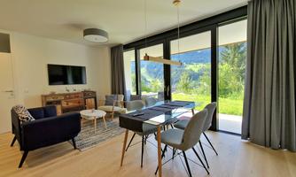 A modern living room with large windows and a view of nature. There is a dining area and cozy seating furniture.