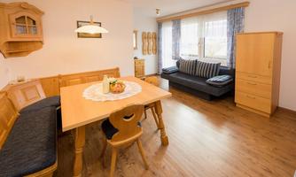 A cozy dining area with a wooden table and a bench. The room is bright and friendly with a sofa and windows that let in plenty of light.