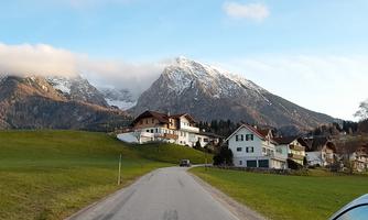 A quiet street winds through a picturesque landscape with houses and tall mountains in the background. The sky is partly cloudy and nature is green and inviting.