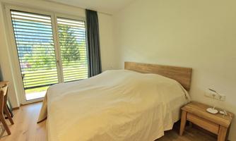 A cozy bedroom with a large bed and a nightstand. The large windows let in plenty of daylight and offer a view of the greenery.