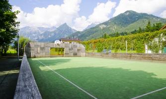 A football field surrounded by mountains and trees. The sky is clear, and the landscape is green and inviting.