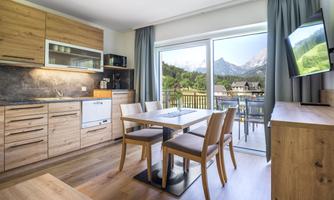 A modern kitchen with wooden interiors and a dining area. Large windows offer a view of the mountains and nature outside.