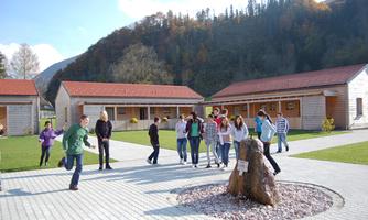 A group of young people is playing and moving on a paved square. In the background, there are modern wooden houses and a wooded mountain landscape.